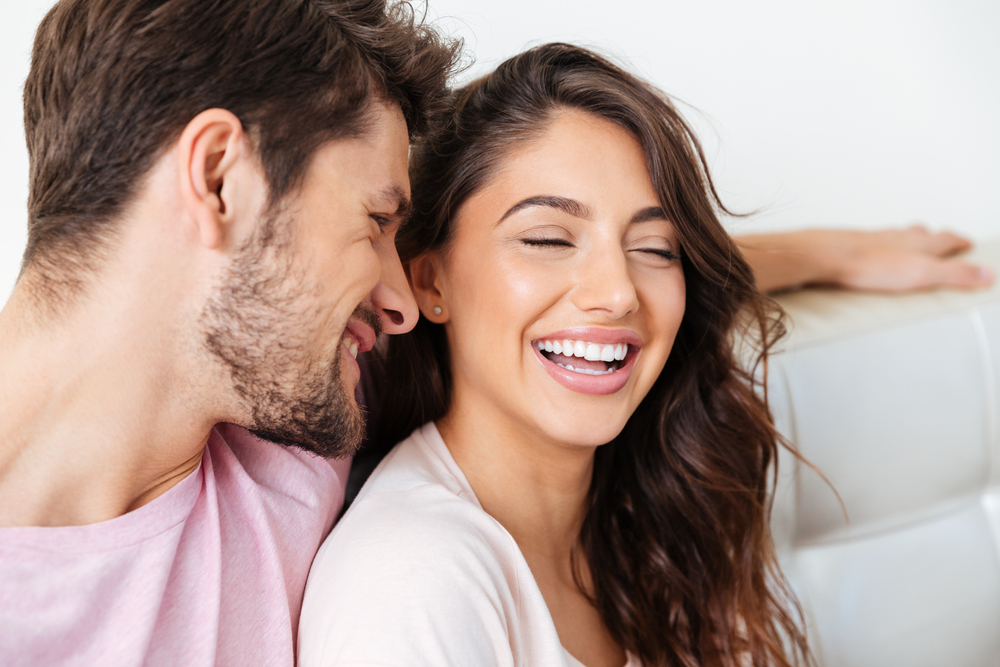 Happy couple laughing together at home - Emergency Dentist in Bonney Lake A joyful couple sharing a lighthearted moment on the couch, showcasing bright and healthy smiles - Emergency Dentist in Bonney Lake