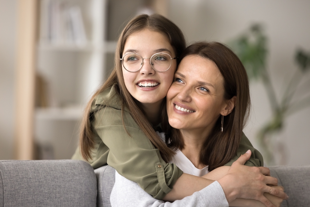 A cheerful daughter embracing her smiling mother while sitting on a couch, showcasing a heartwarming family connection - Bellevue Dentist