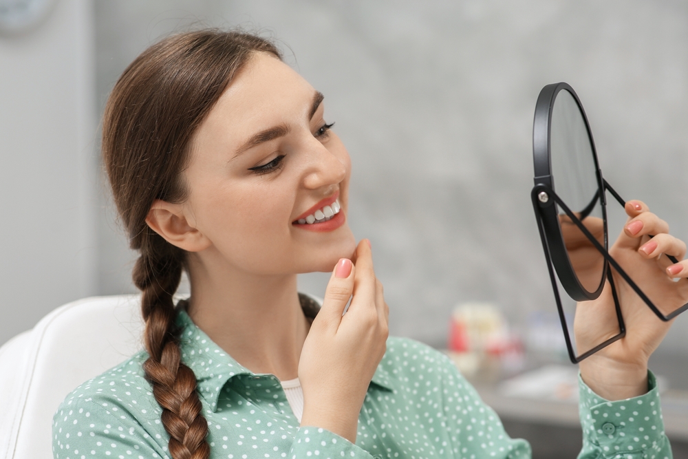 Woman admiring her smile in mirror at dentist - Emergency Dentist Sumner A young woman checking her teeth in a mirror while smiling after a dental appointment - Emergency Dentist Sumner
