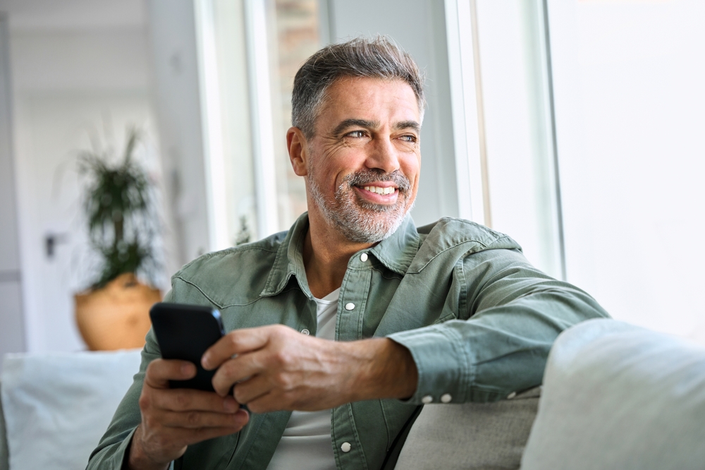 Mature man smiling while holding phone - Emergency Dentist Sumner A relaxed middle-aged man with a beard smiling as he sits indoors holding his smartphone - Emergency Dentist Sumner