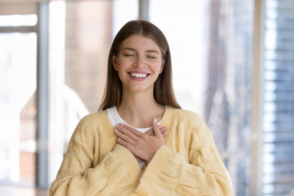 Woman smiling with gratitude and happiness - Bellevue Dentist A young woman with eyes closed and hands on her chest, smiling warmly and radiating joy - Bellevue Dentist