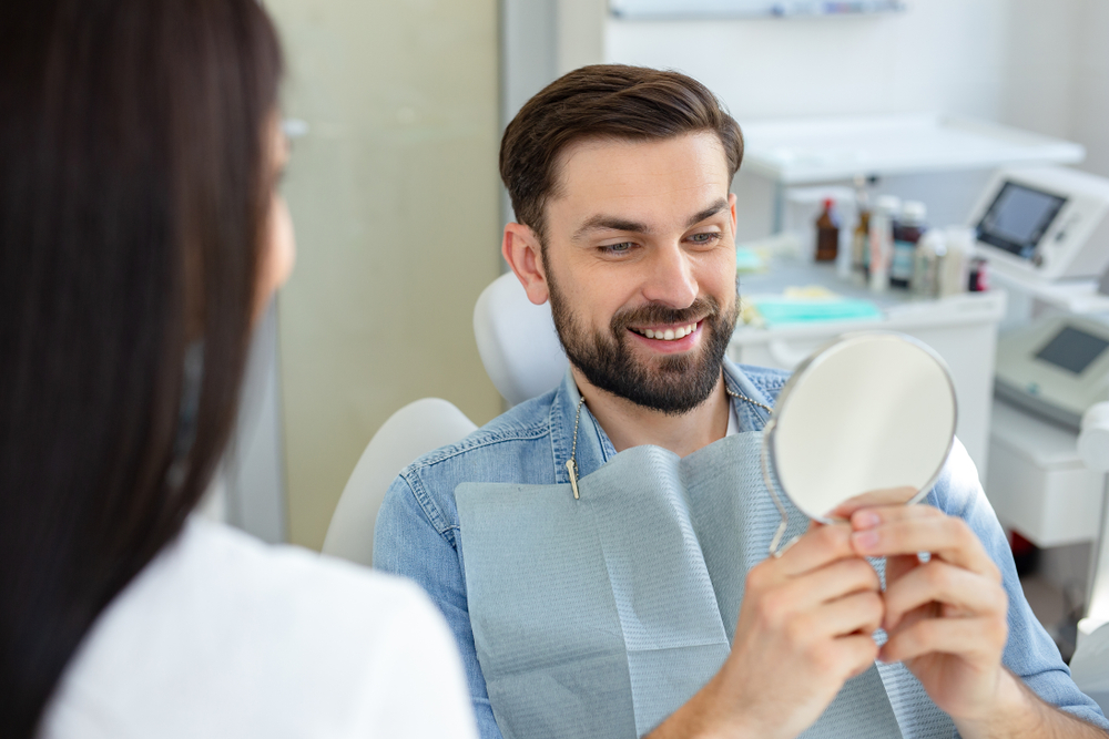 A man sitting in a dental chair happily examining his teeth in a handheld mirror after treatment - Emergency Dentist in DuPont
