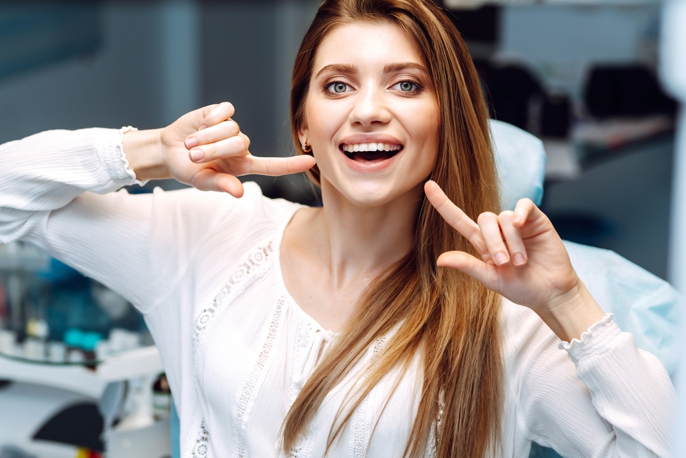 Young woman showing her smile at the dental clinic - Emergency Dentist in Bonney Lake A cheerful woman sitting in a dental chair confidently pointing to her bright smile after treatment - Emergency Dentist in Bonney Lake