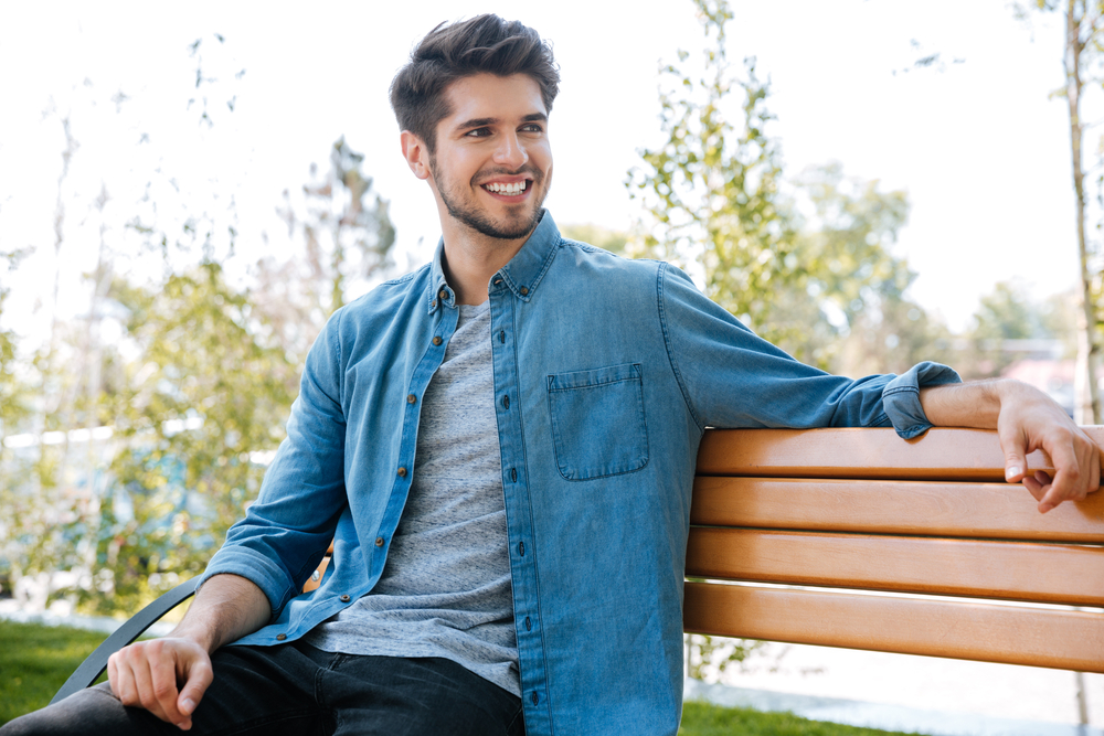 Young man smiling on a park bench - Bellevue Dentist A cheerful young man in casual clothes sitting on a bench outdoors, enjoying a bright day with a confident smile - Bellevue Dentist